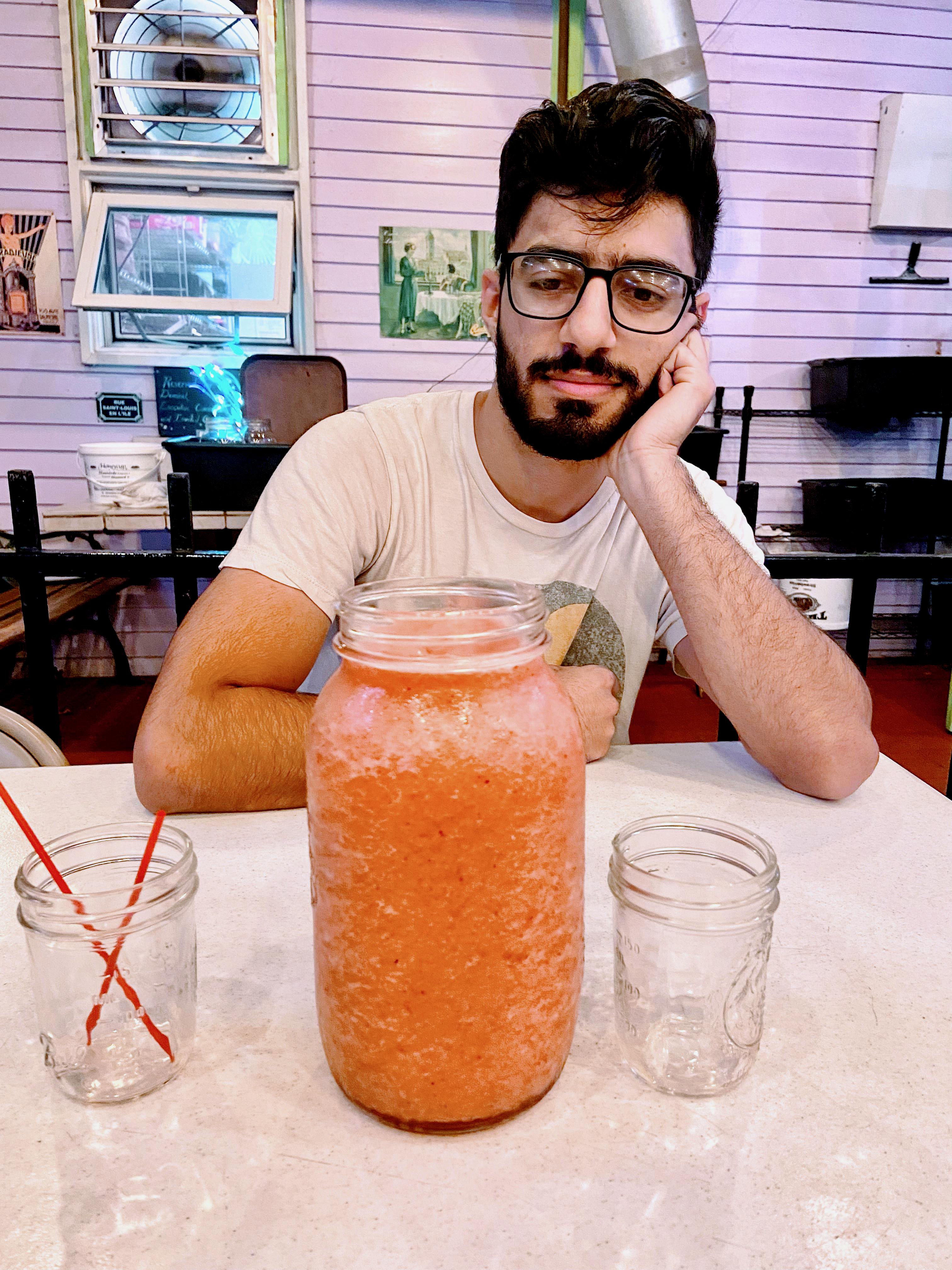 A boy staring at a large jar of a light pink smoothie. He is wearing glasses.